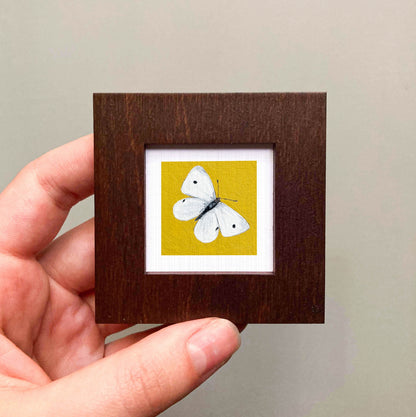 A hand is holding a small wooden frame containing a framed image of a white butterfly against a yellow background.