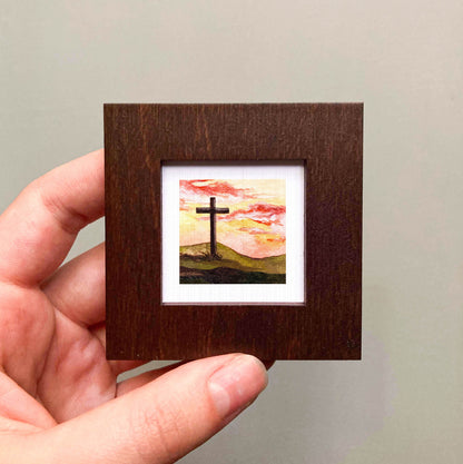 A hand is holding a small wooden frame containing a painting of a cross against a sunset landscape.