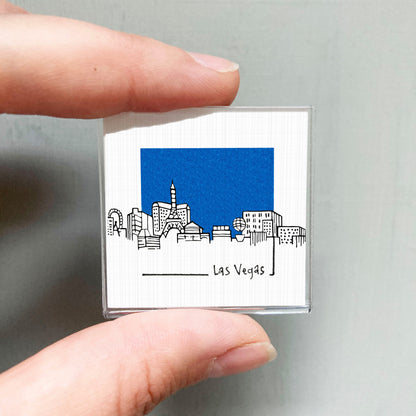 A person's hand holding a small square magnet with a cityscape illustration on it, featuring the text "Las Vegas" below the image.