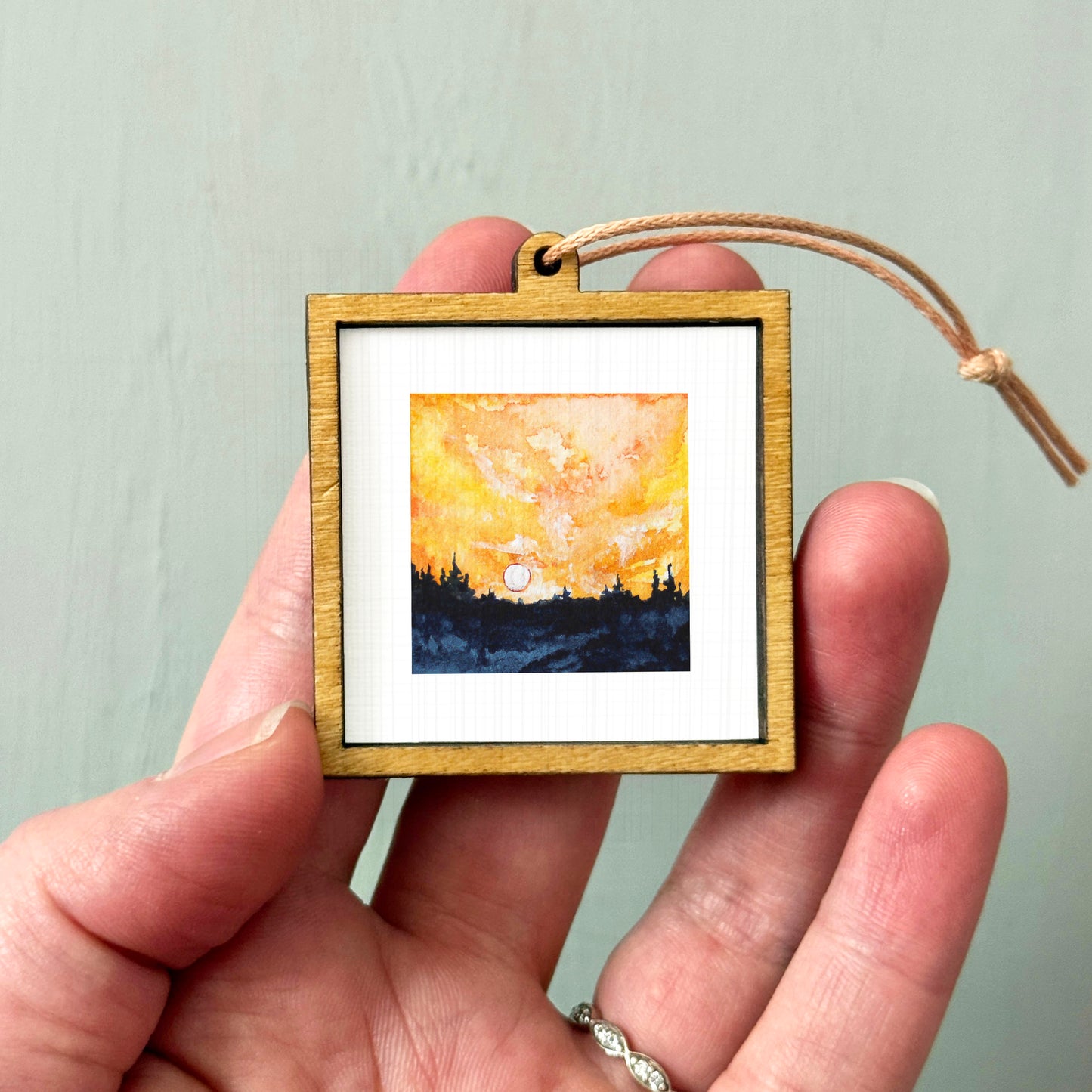 A hand holding a small wooden frame containing a painting of a sunset over a landscape.