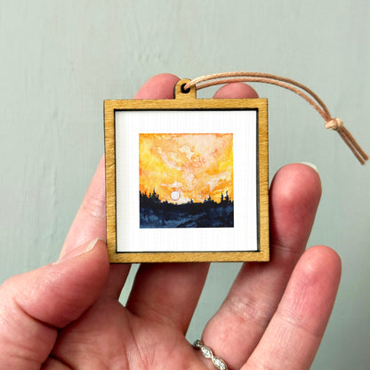 A hand holding a small wooden frame containing a painting of a sunset over a landscape.