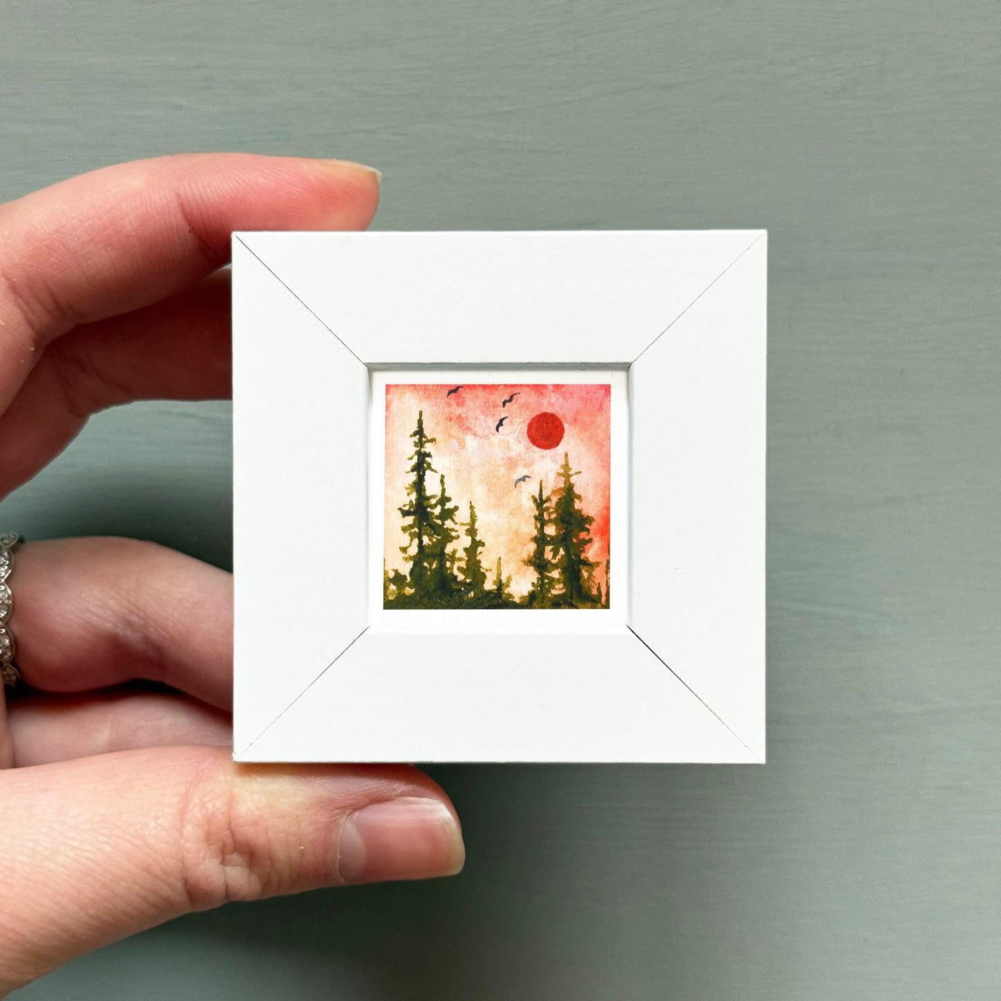A hand is holding a small white square frame containing a painting of a forest scene with a red sun in the sky.
