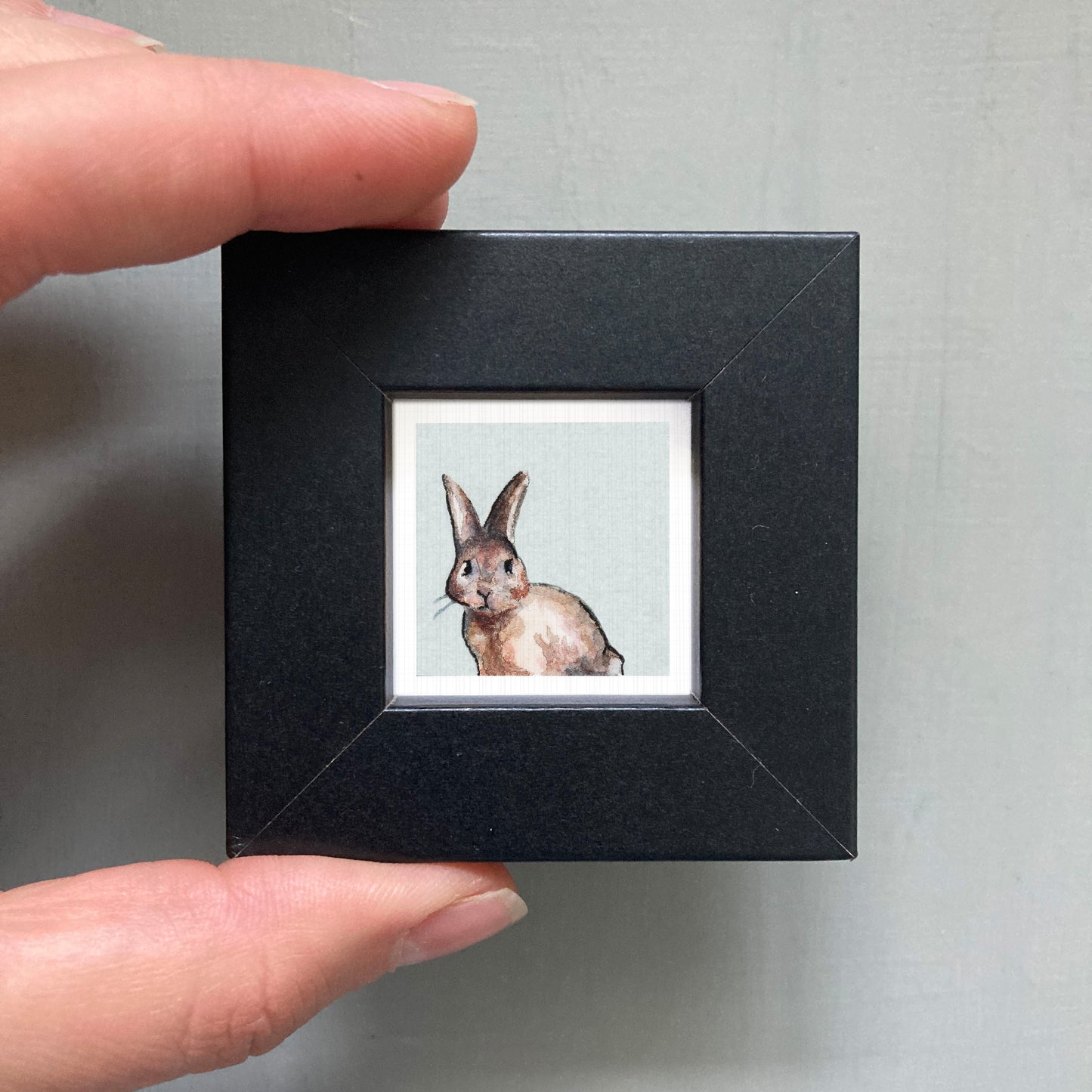 A person's hand holding a small black picture frame containing a painting of a rabbit.