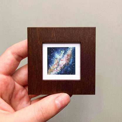 A hand is holding a small wooden frame containing a miniature painting of a galaxy.