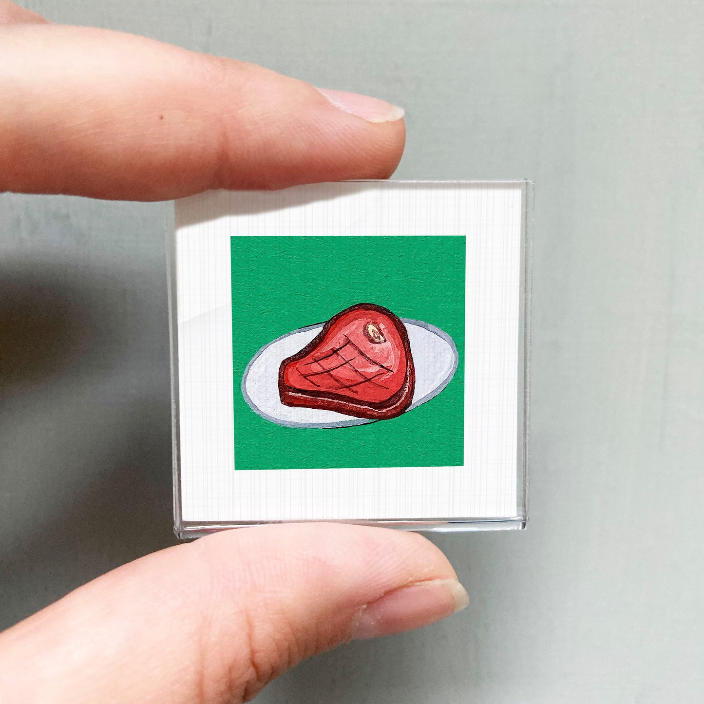 A hand is holding a small square with a red meat illustration on a green background.