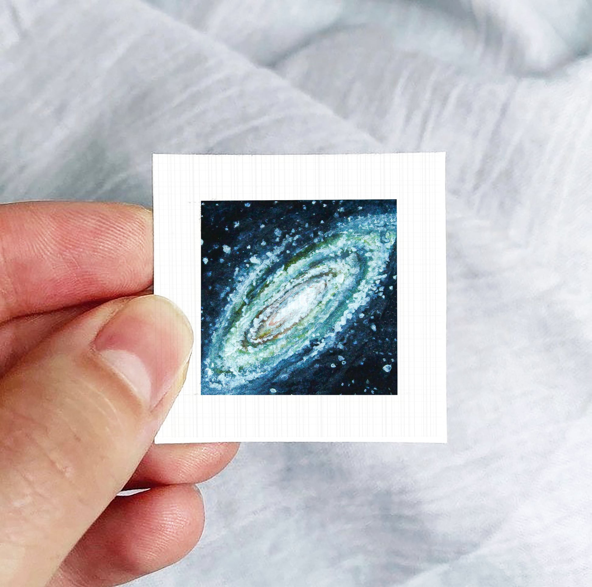 A hand is holding a small square photograph of a spiral galaxy against a white background.