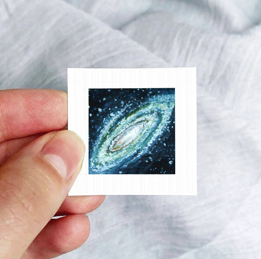 A hand is holding a small square photograph of a spiral galaxy against a white background.