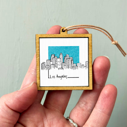 A hand holding a small wooden frame with a picture of a city skyline and the text "Los Angeles" written on it.