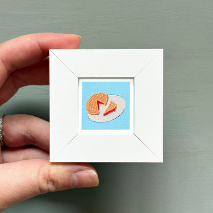 A hand is holding a small white frame containing an embroidered image of a piece of food, likely a pancake or waffle, on a blue background.
