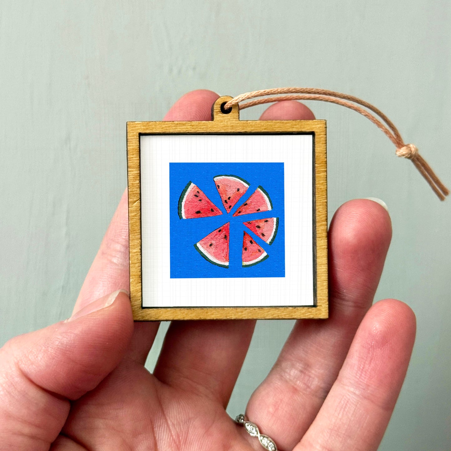 A hand holding a small wooden frame containing a framed image of watermelon slices.