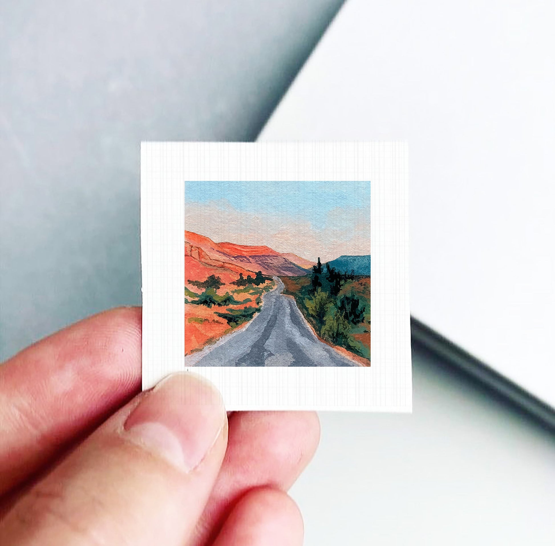 A hand is holding a small square-shaped piece of paper with a painting of a winding road through a desert landscape.