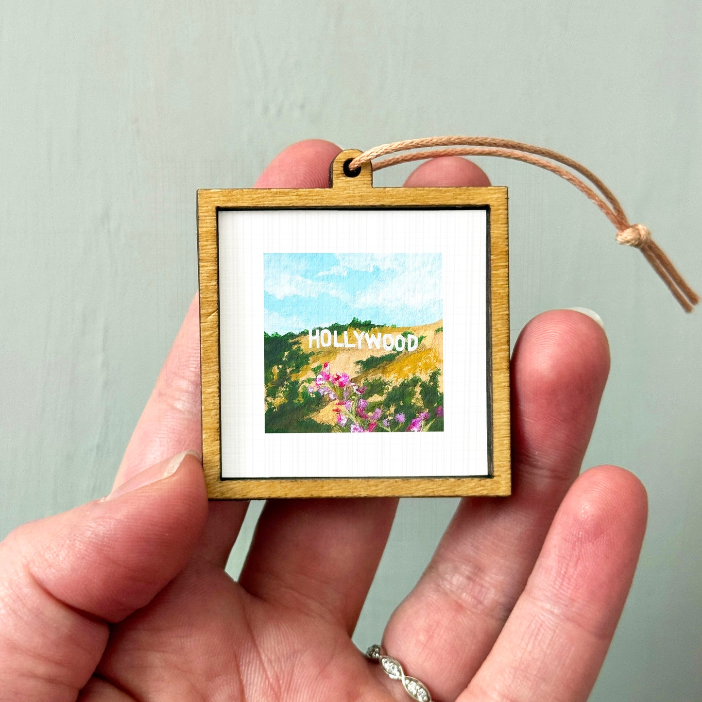 A person's hand holding a small wooden frame containing a framed image of a scenic landscape with mountains and flowers.
