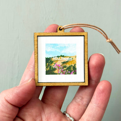 A person's hand holding a small wooden frame containing a framed image of a scenic landscape with mountains and flowers.