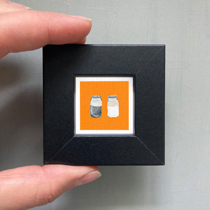A hand is holding a small black frame containing an illustration of two salt and pepper shakers.