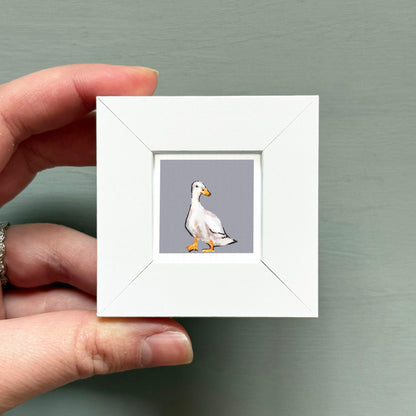 A hand is holding a small white square frame containing a single image of a duck.