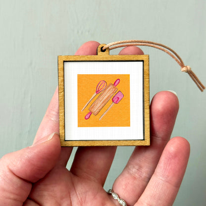 A hand holding a small wooden frame containing an illustration of a rolling pin and a wooden spoon.