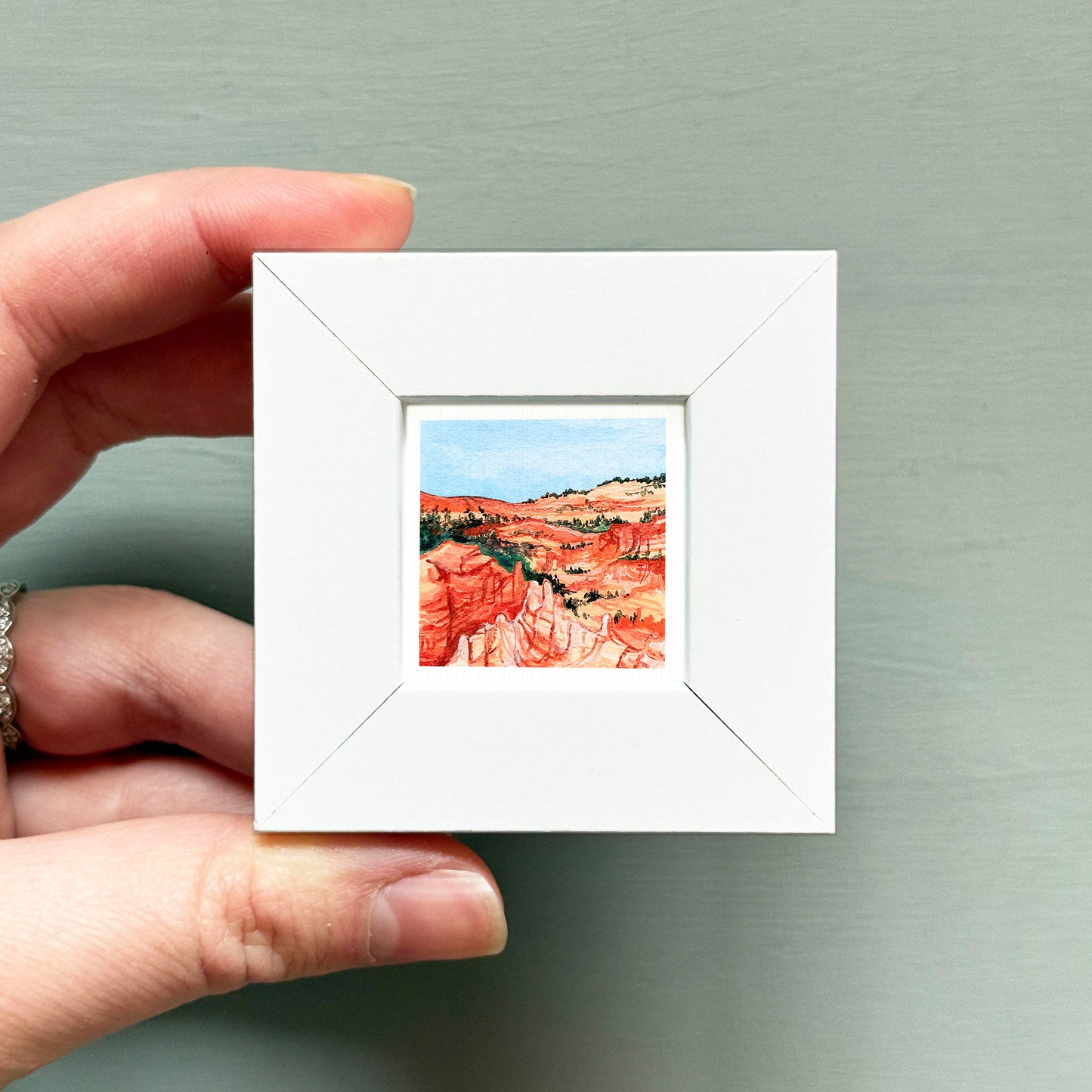 A hand is holding a small white square frame containing a watercolor painting of a desert landscape.