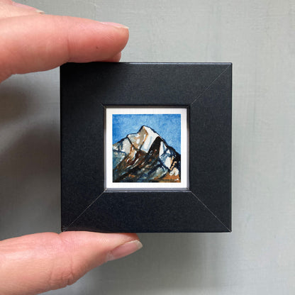 A person's hand holding a small black frame containing a framed painting of a mountain landscape.