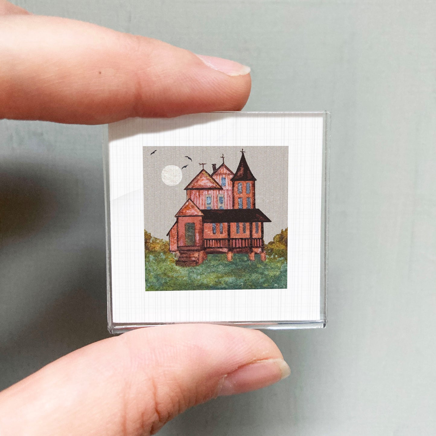 A hand holding a small square picture of a house with a red roof and a white moon in the sky.