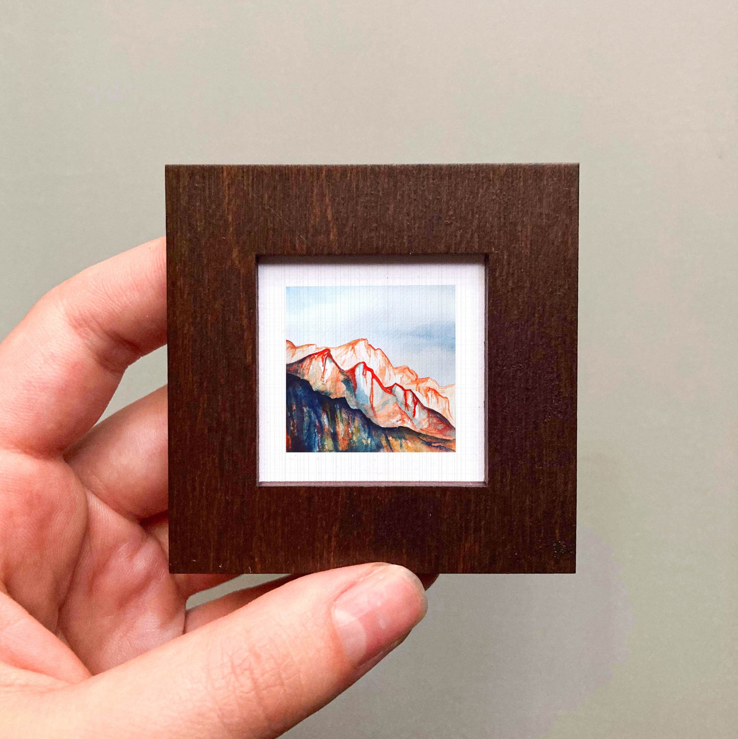 A hand is holding a small, square-shaped wooden frame containing a miniature painting of a mountain landscape.