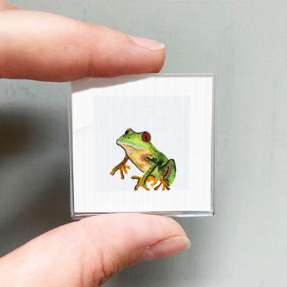 A person's hand is holding a small square with a detailed illustration of a green frog on it.