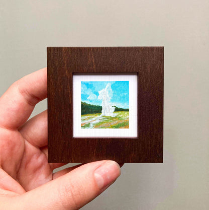 A hand is holding a small square picture frame containing a painting of a landscape with a fountain in the background.