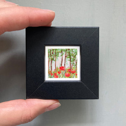 A hand is holding a small black frame containing a framed painting of red flowers in a forest.
