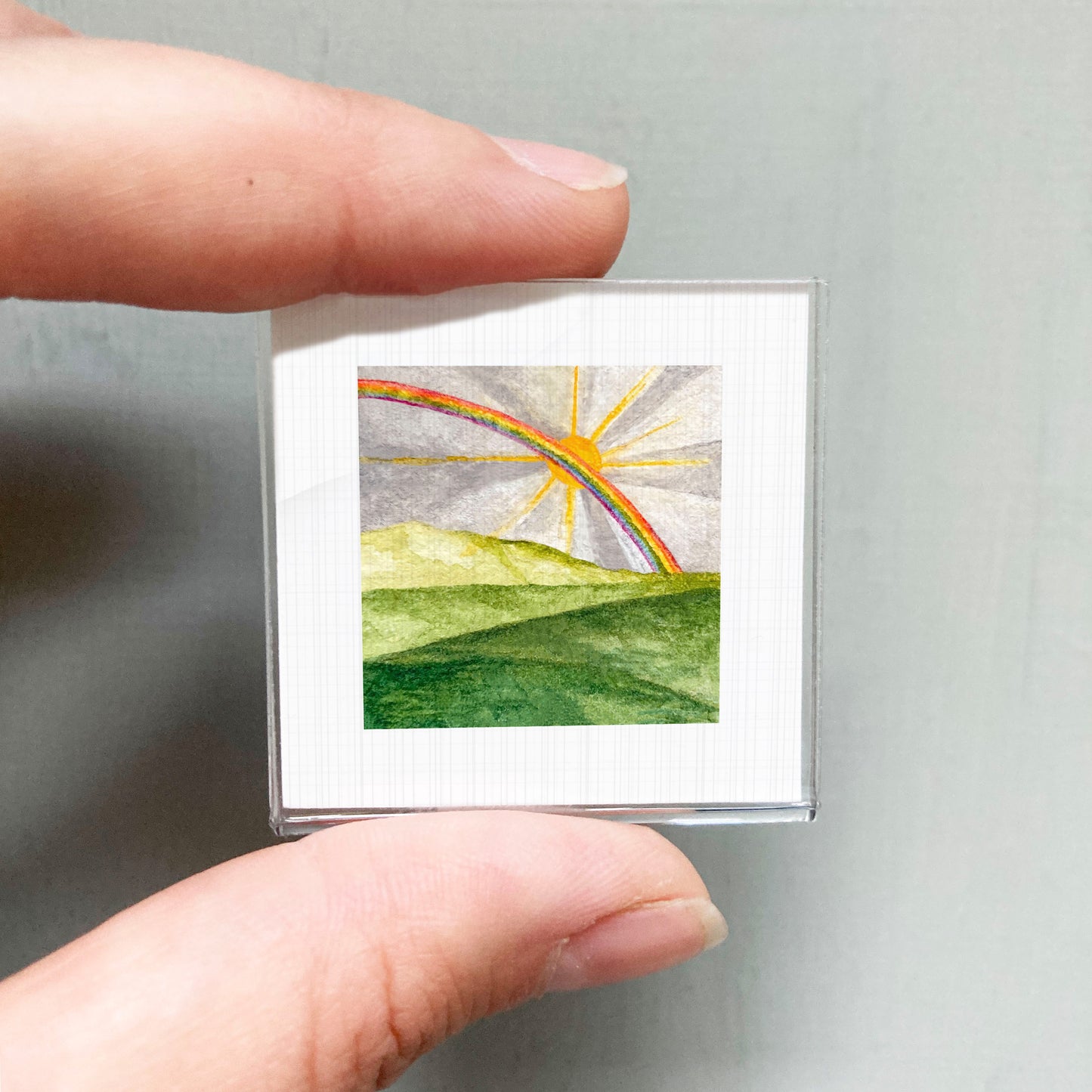 A person's hand is holding a small square picture of a rainbow against a green landscape.