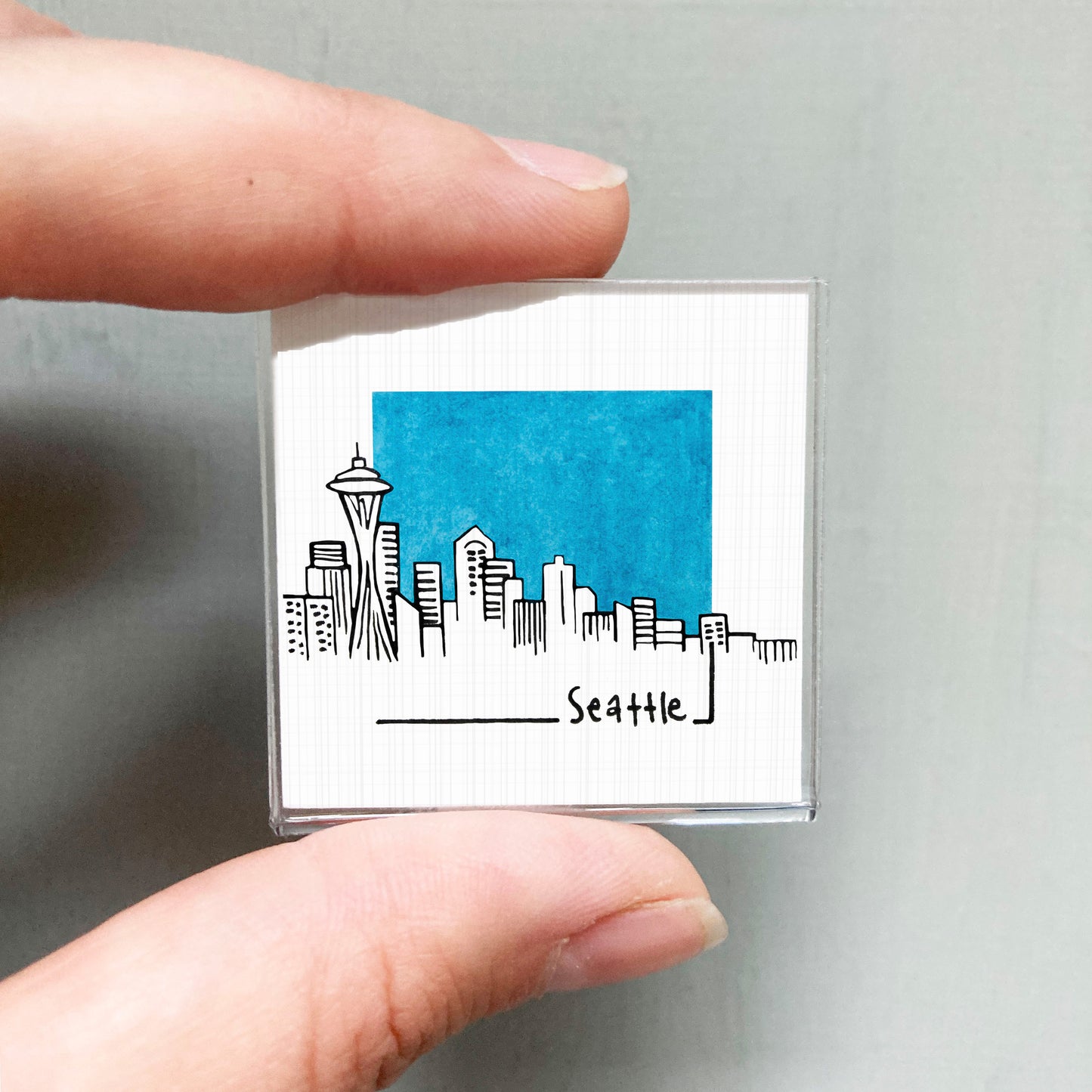 A hand holding a small square magnet with a drawing of the Seattle skyline and the word "Seattle" written below it.