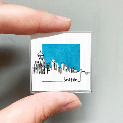 A hand holding a small square magnet with a drawing of the Seattle skyline and the word "Seattle" written below it.