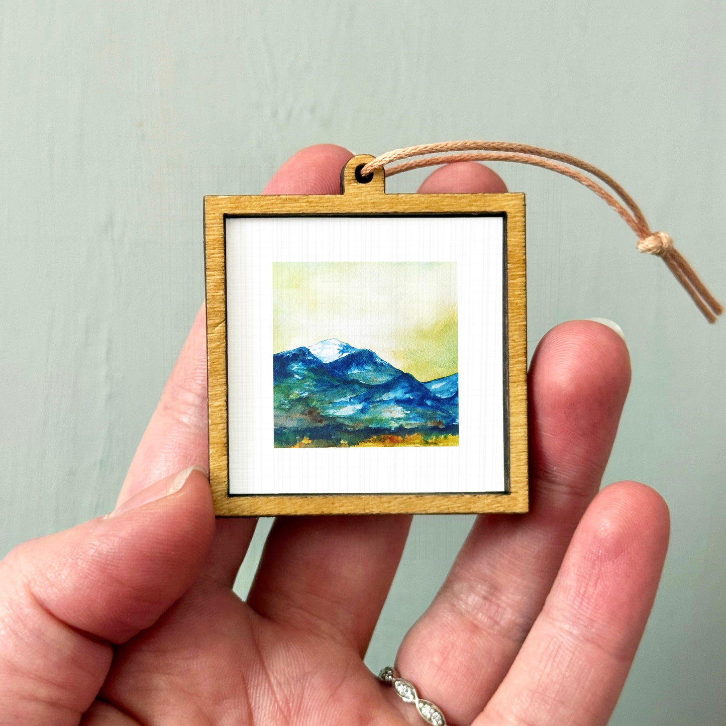 A hand holding a small wooden frame containing a watercolor painting of a mountain landscape.