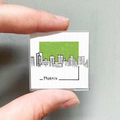 A person's hand holding a small square magnet with a cityscape illustration on it.