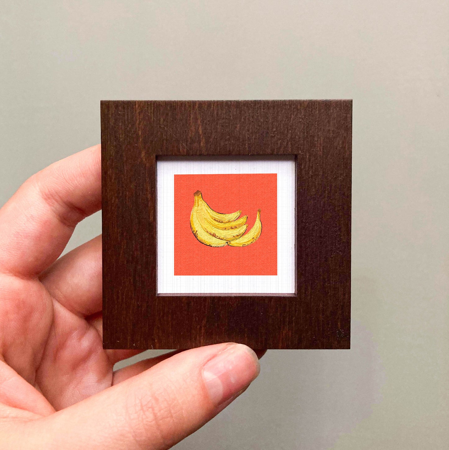 A hand is holding a small wooden frame containing a framed image of a bunch of bananas.