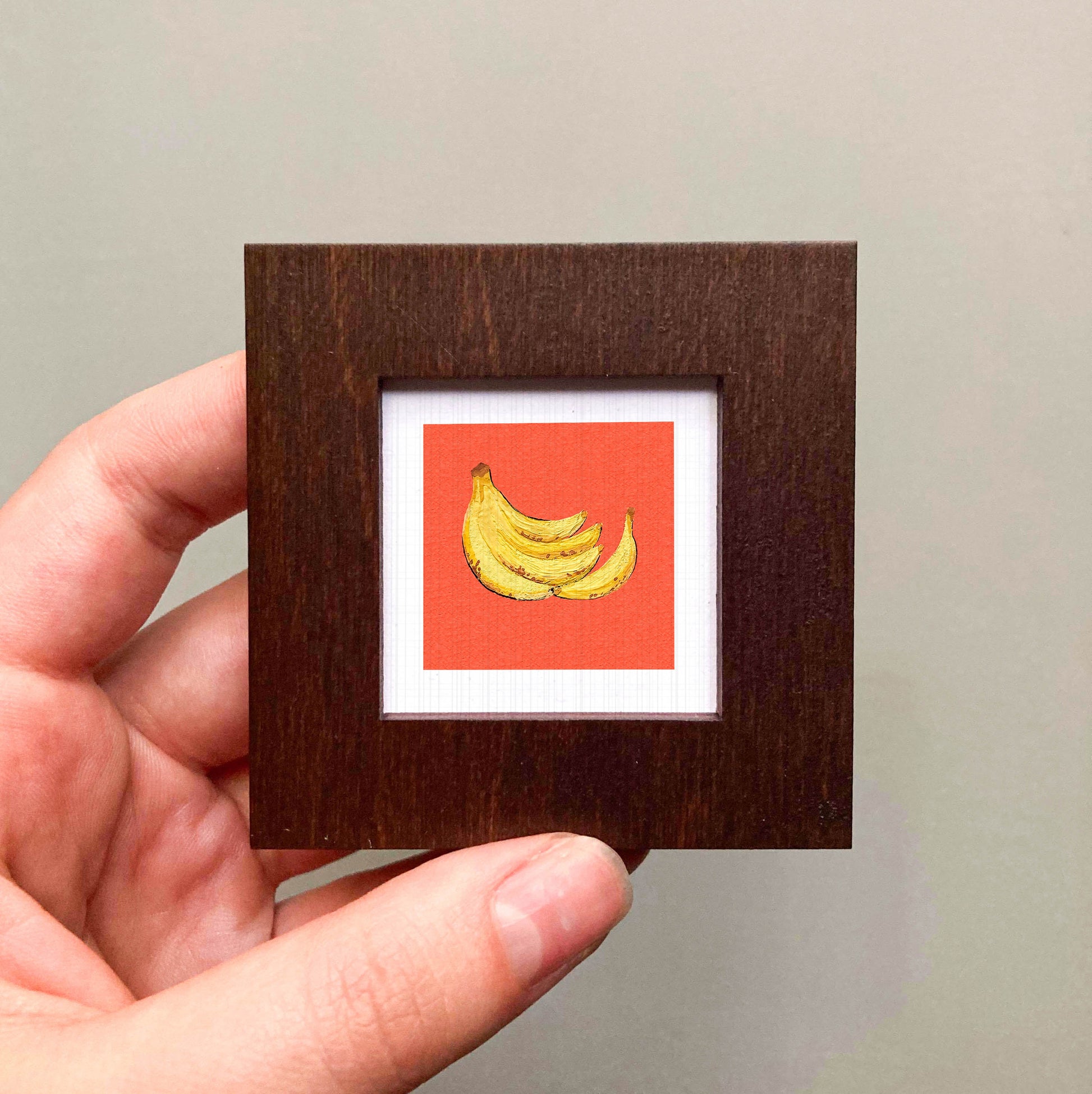 A hand is holding a small wooden frame containing a framed image of a bunch of bananas.