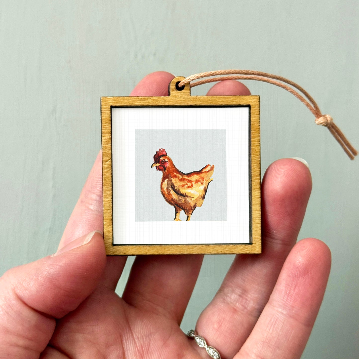 A person's hand holding a small wooden frame containing a picture of a chicken.
