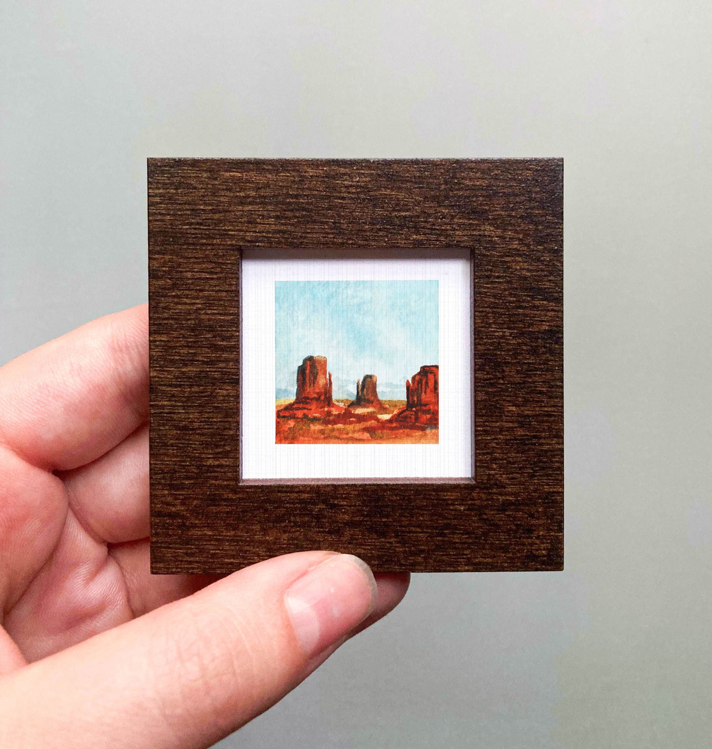 A hand is holding a small framed picture of a desert landscape with rock formations.