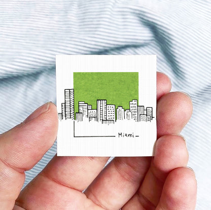 A hand is holding a small square-shaped paper with a drawing of a city skyline and the word " Miami" written on it.