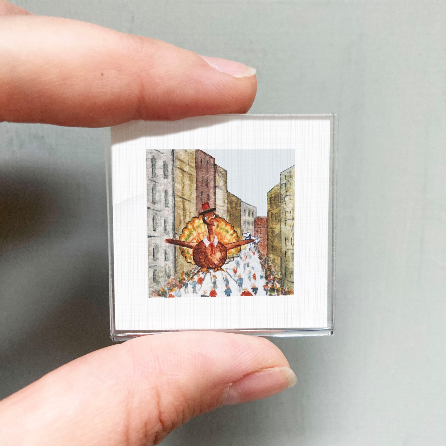 A person's hand is holding a small square picture depicting a turkey in a city street scene.