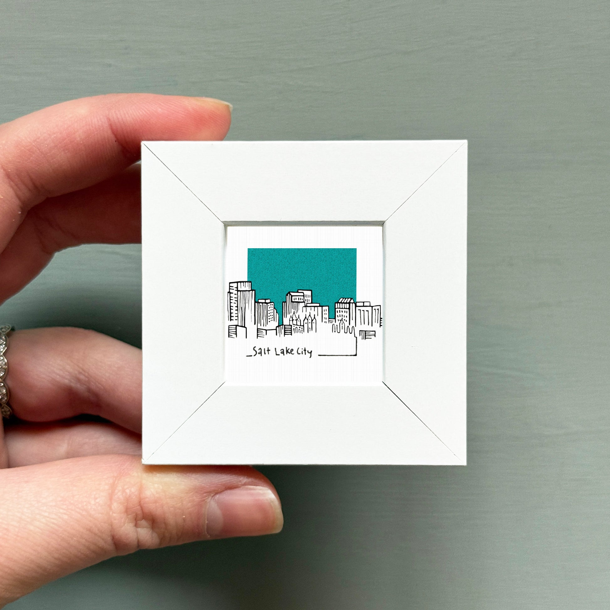 A hand is holding a small white square frame containing a drawing of a city skyline.