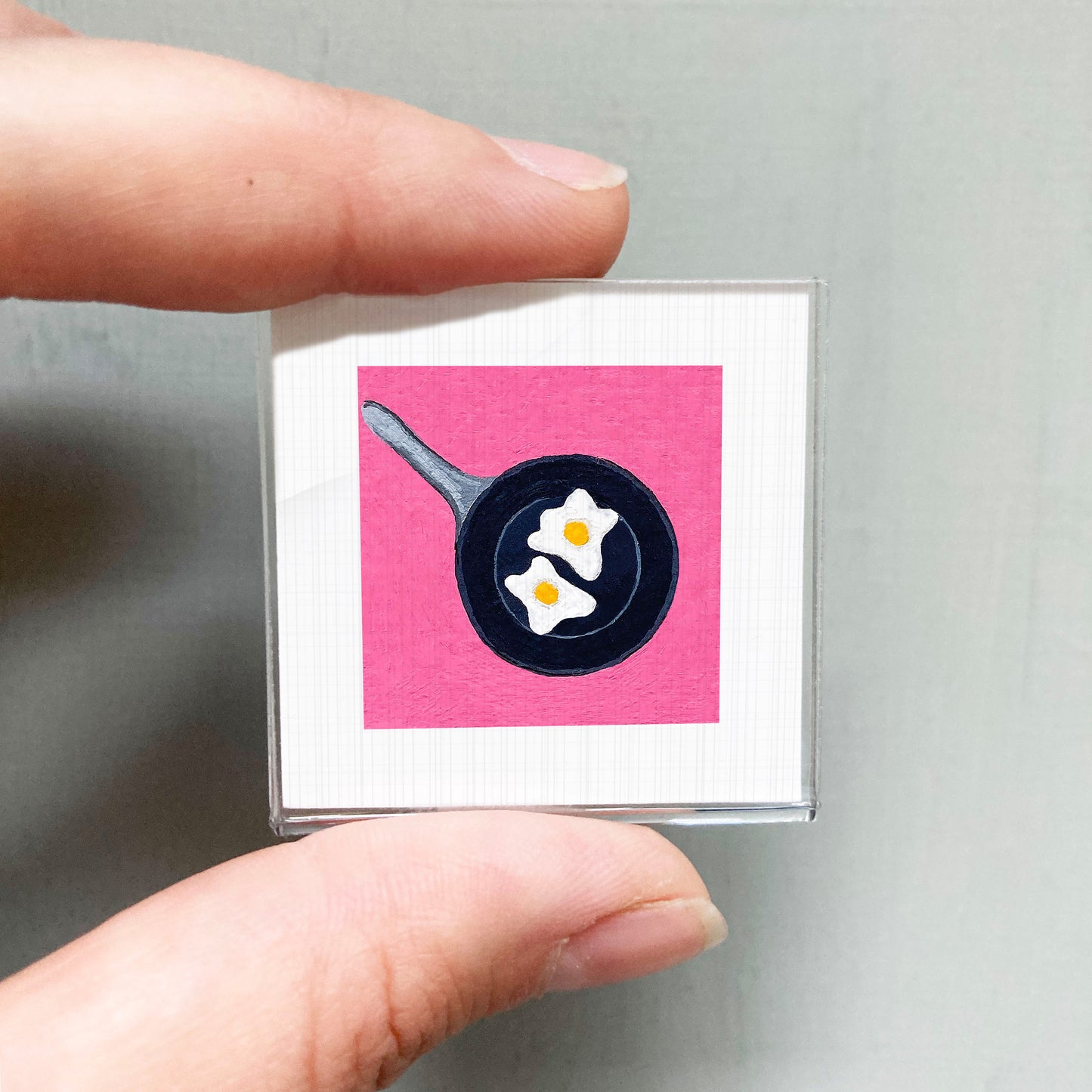 A person's hand holding a small square with a pink background and a black frying pan with two white eggs on it.