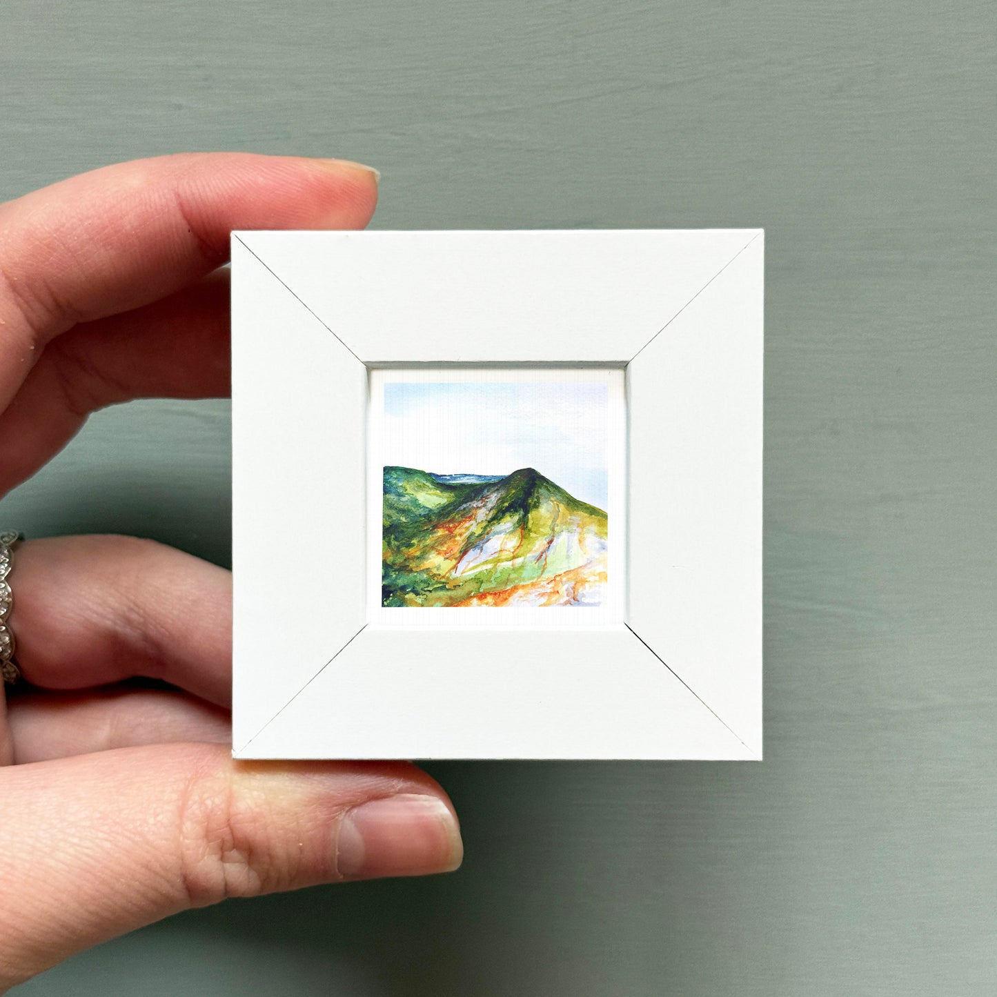 A hand is holding a small white picture frame containing a landscape painting of a mountain.