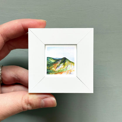 A hand is holding a small white picture frame containing a landscape painting of a mountain.
