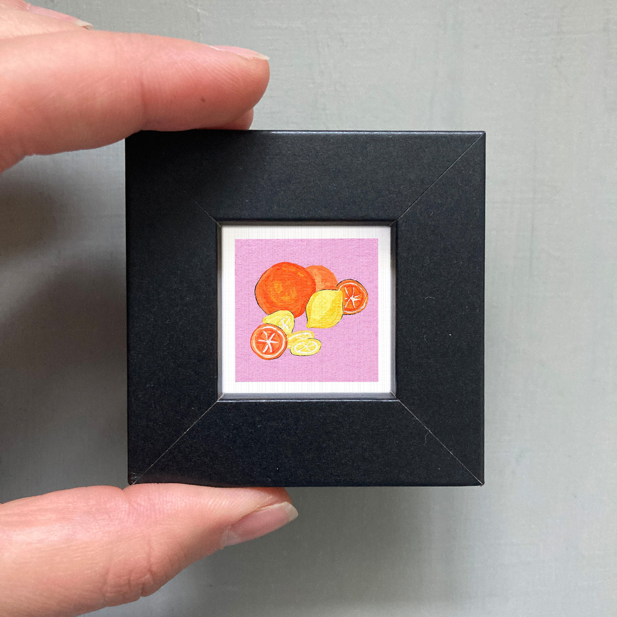 A hand is holding a small square picture frame containing an illustration of oranges and lemons.