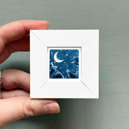 A hand is holding a small white square frame containing a painting of a crescent moon and stars against a dark blue night sky.