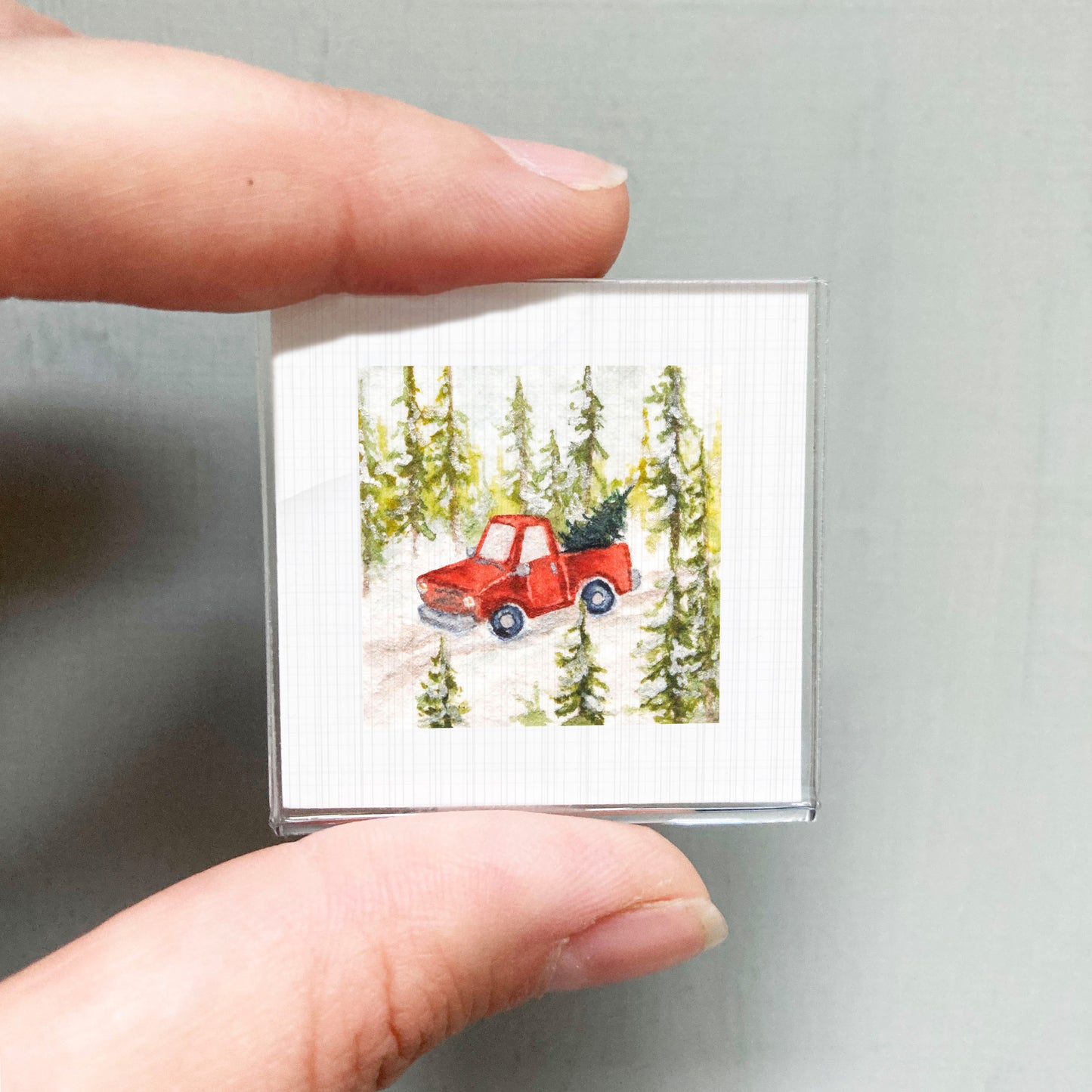 A person's hand is holding a small square picture depicting a red truck driving through a snowy forest with pine trees.