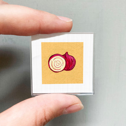 A person's hand holding a small square with an illustration of a red onion on it.