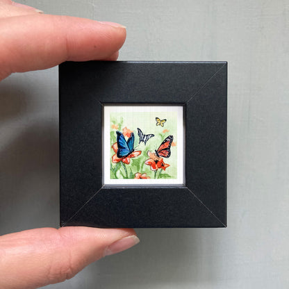 A person's hand is holding a small black frame containing a framed painting of three butterflies on flowers.