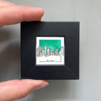 A hand is holding a small black square frame containing a drawing of a city skyline with green buildings.