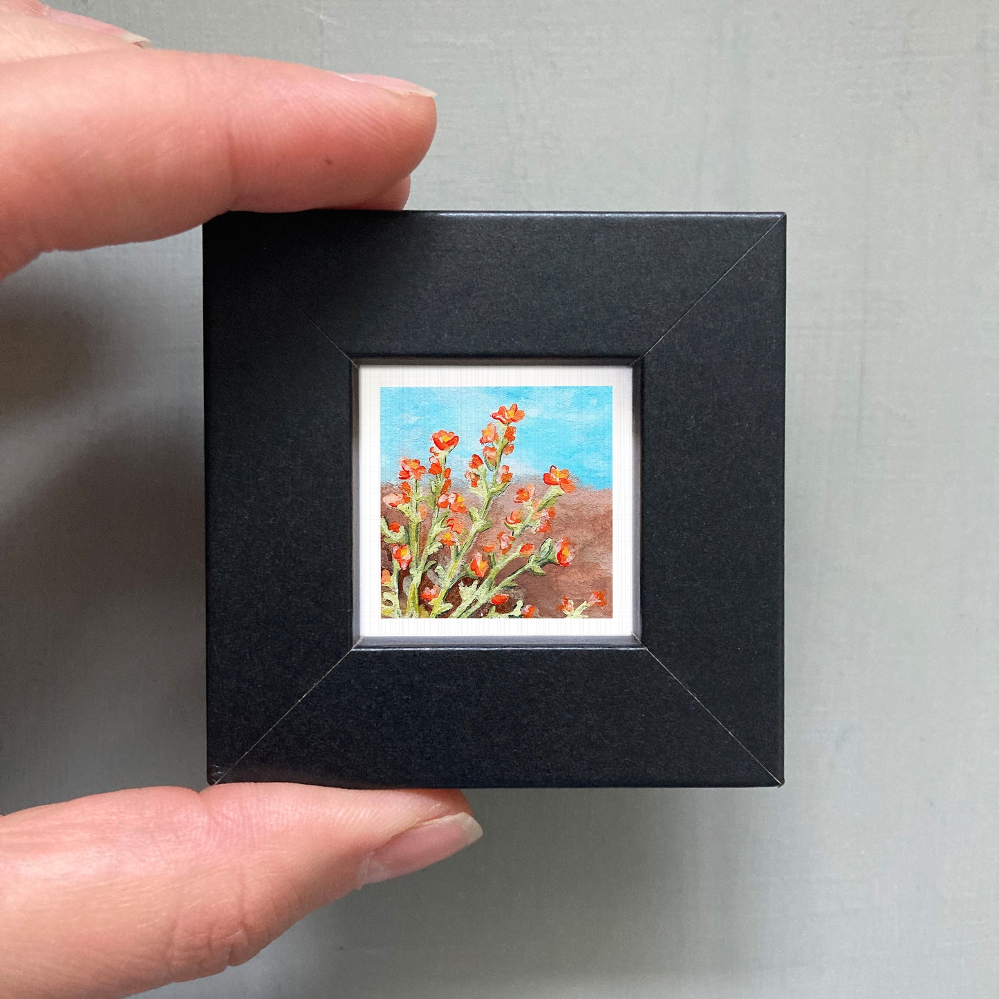A person's hand holding a small black picture frame containing a painting of red flowers against a blue background.