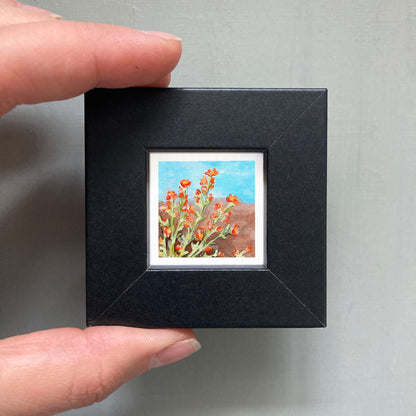 A person's hand holding a small black picture frame containing a painting of red flowers against a blue background.